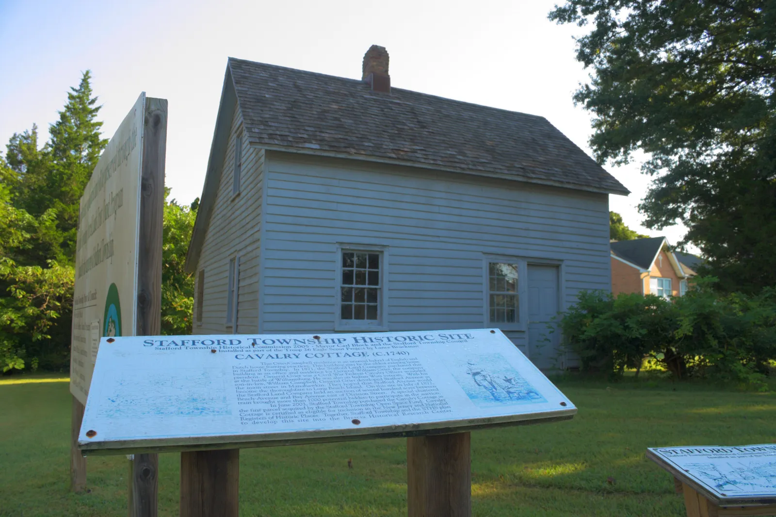 A small pale gray clapboard cottage behind a historic site marker on the lawn