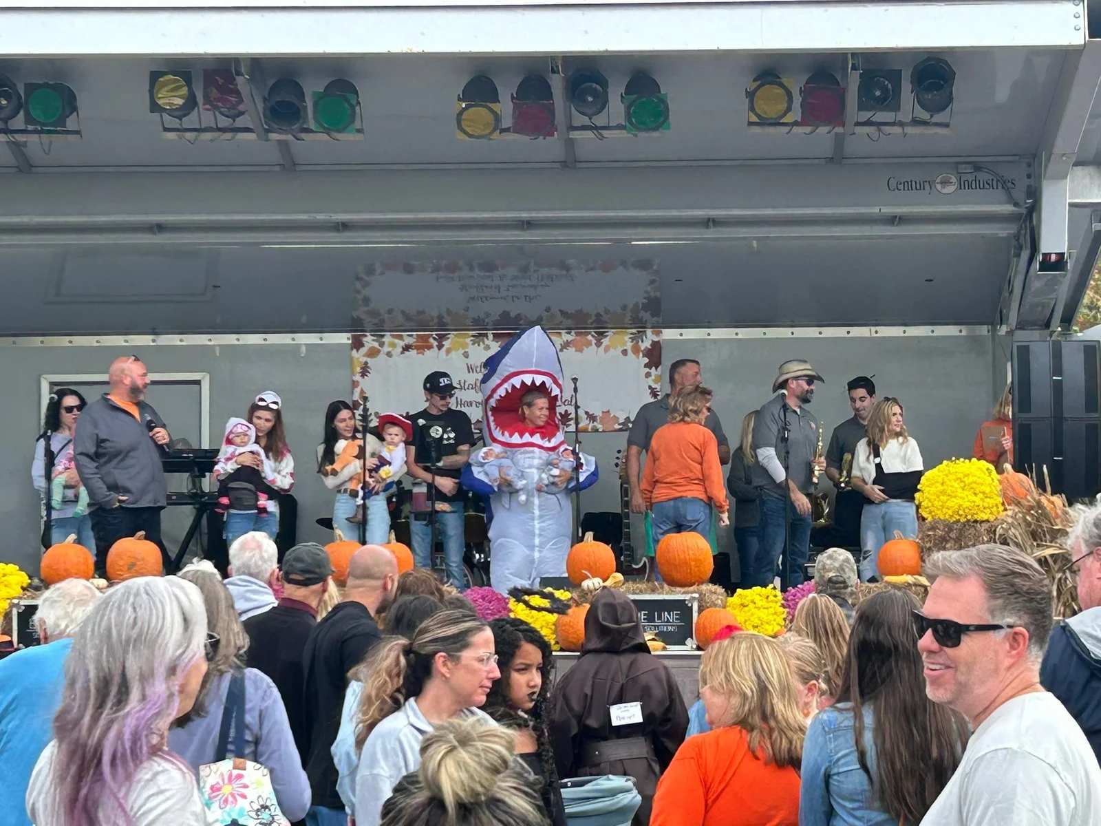 A crowd gathered at an outdoor stage decorated with pumpkins and mums, with costumed participants on stage, at Stafford's Fall Harvest Festival