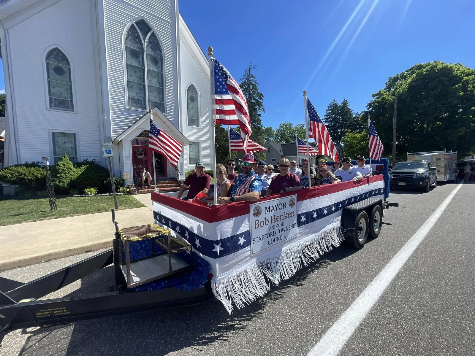 A red, white and blue parade float with American flags passing a white church during Stafford's Founders Day parade