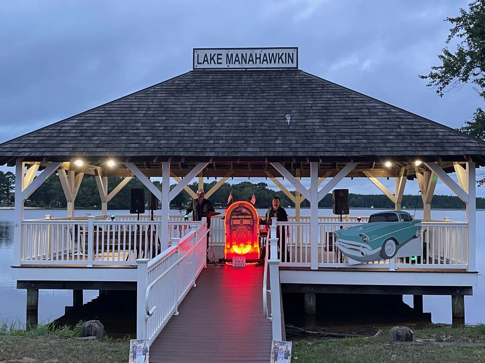 The Lake Manahawkin gazebo lit up at twilight for a summer concert, with a vintage car cutout and a glowing jukebox on stage