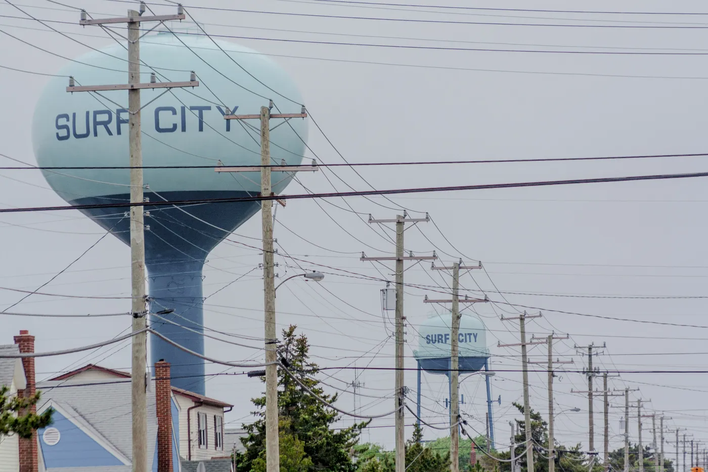 A pale blue water tower painted Surf City, with a second tower and a line of utility poles behind it