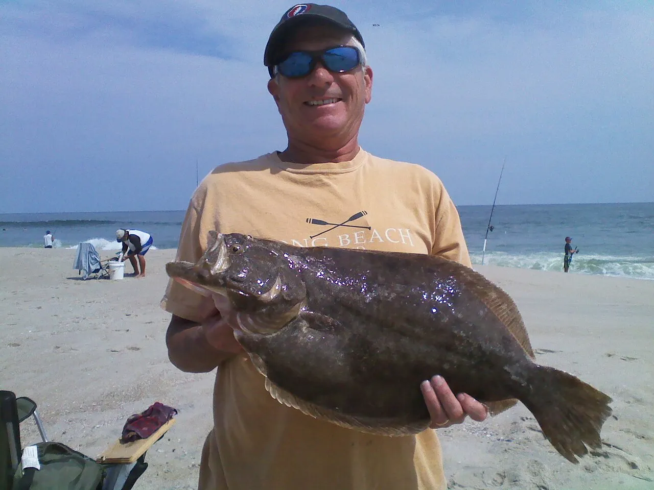 A smiling angler on the beach holding up a large flounder, with fishing rods and the ocean behind him