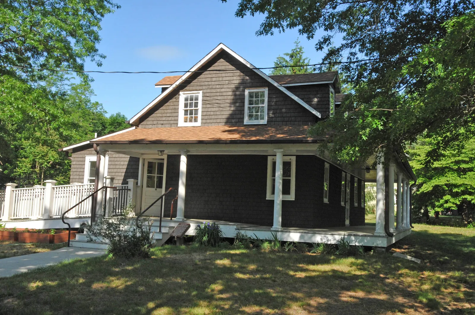 A brown shingled 19th-century house with a wraparound porch, surrounded by trees