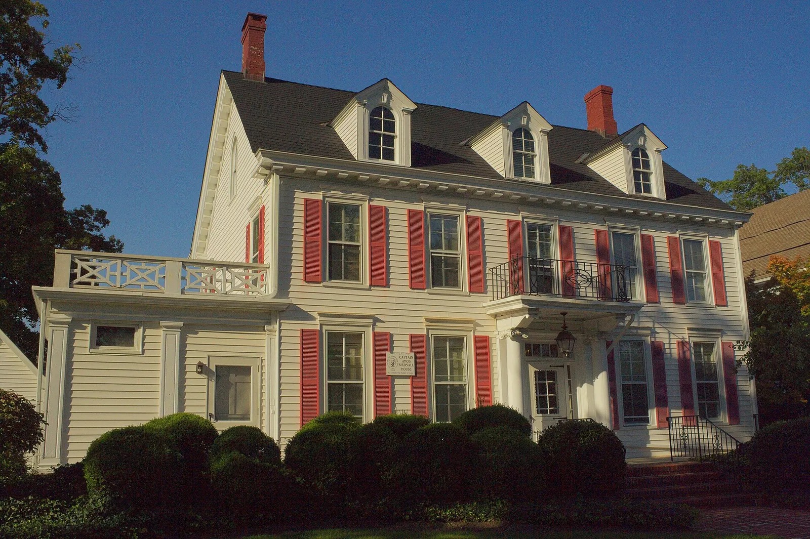 Two-story Colonial-style house with red shutters and dormers behind green shrubs