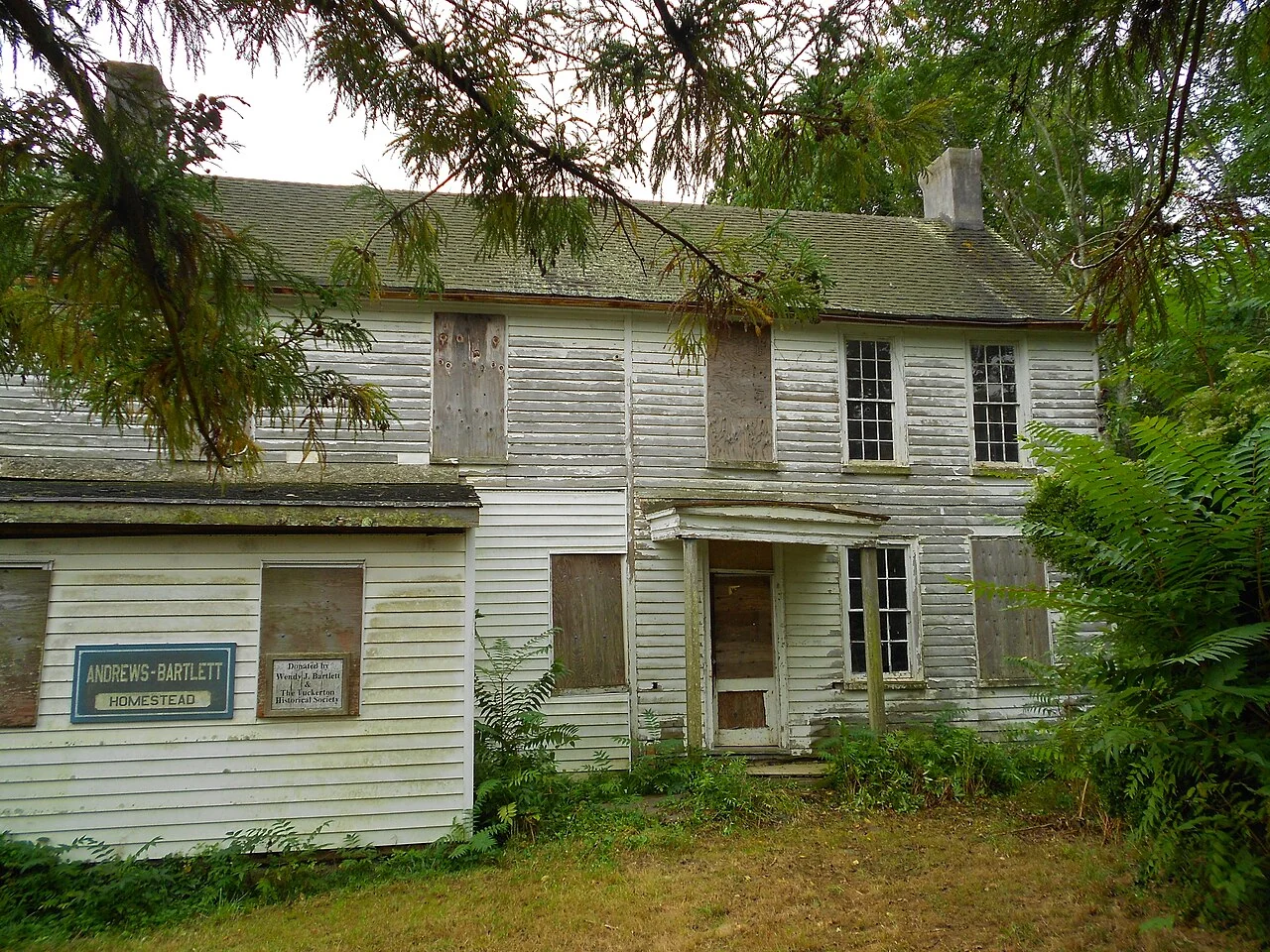 The weathered white clapboard Andrews-Bartlett Homestead with its sign, framed by trees and ferns