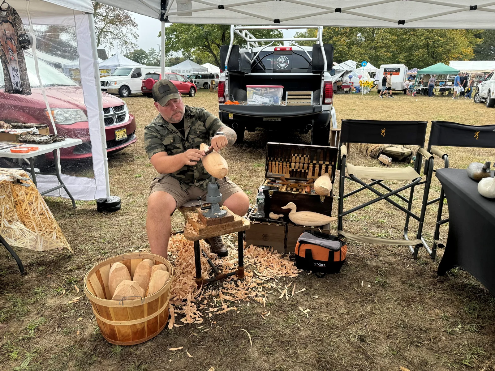 A carver shaping a wooden decoy at his booth, with a basket of carved blanks and wood shavings at his feet and vendor tents behind