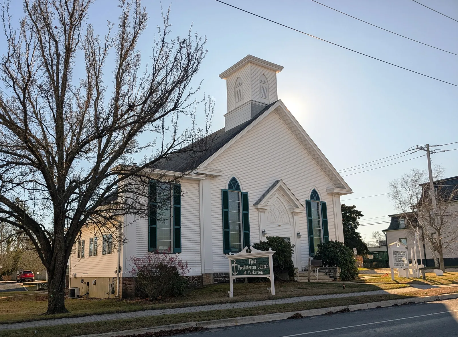 A white clapboard church with green shutters and a cupola beside a large bare tree