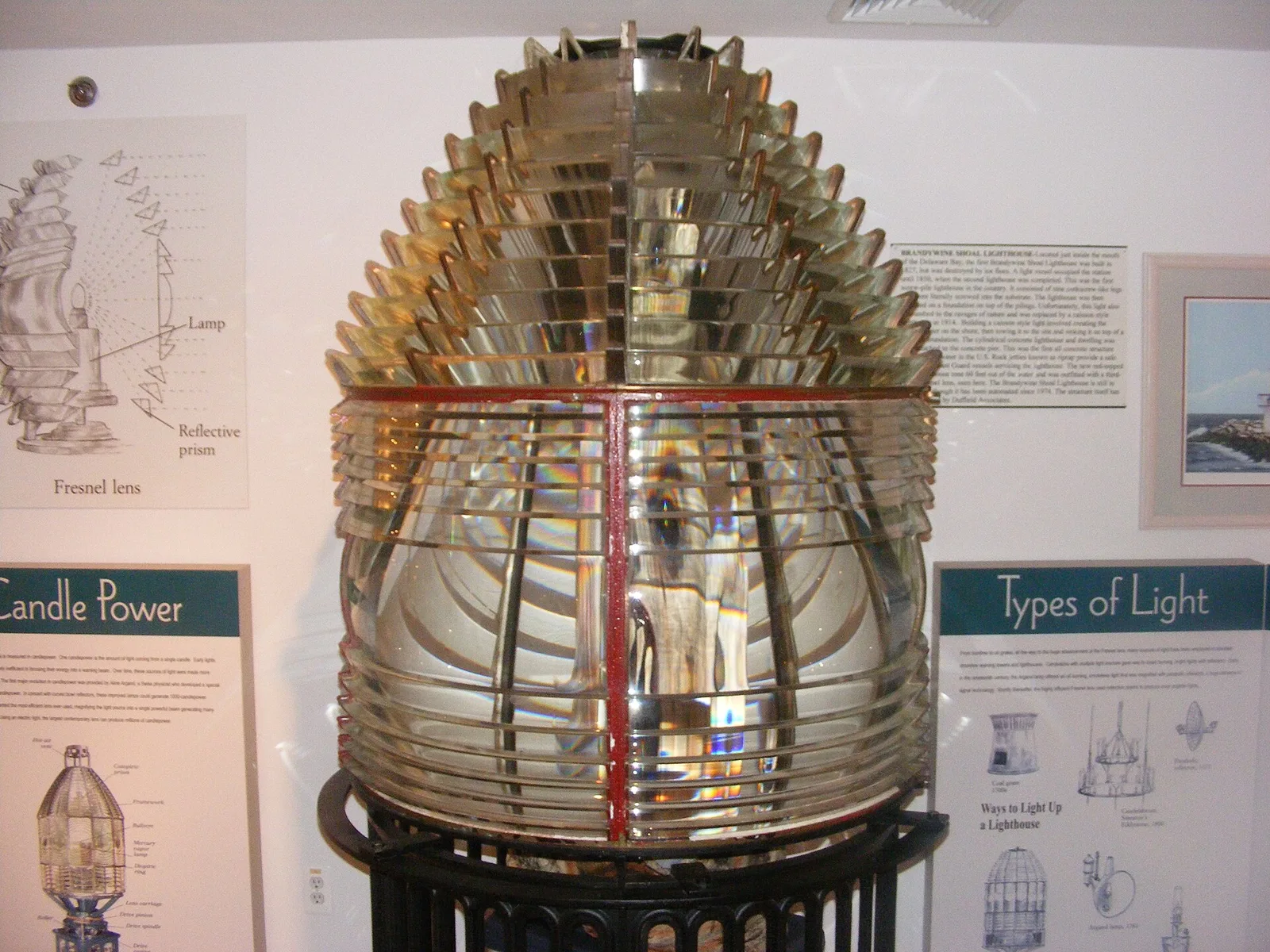 A tall glass Fresnel lighthouse lens on display with exhibit panels behind it