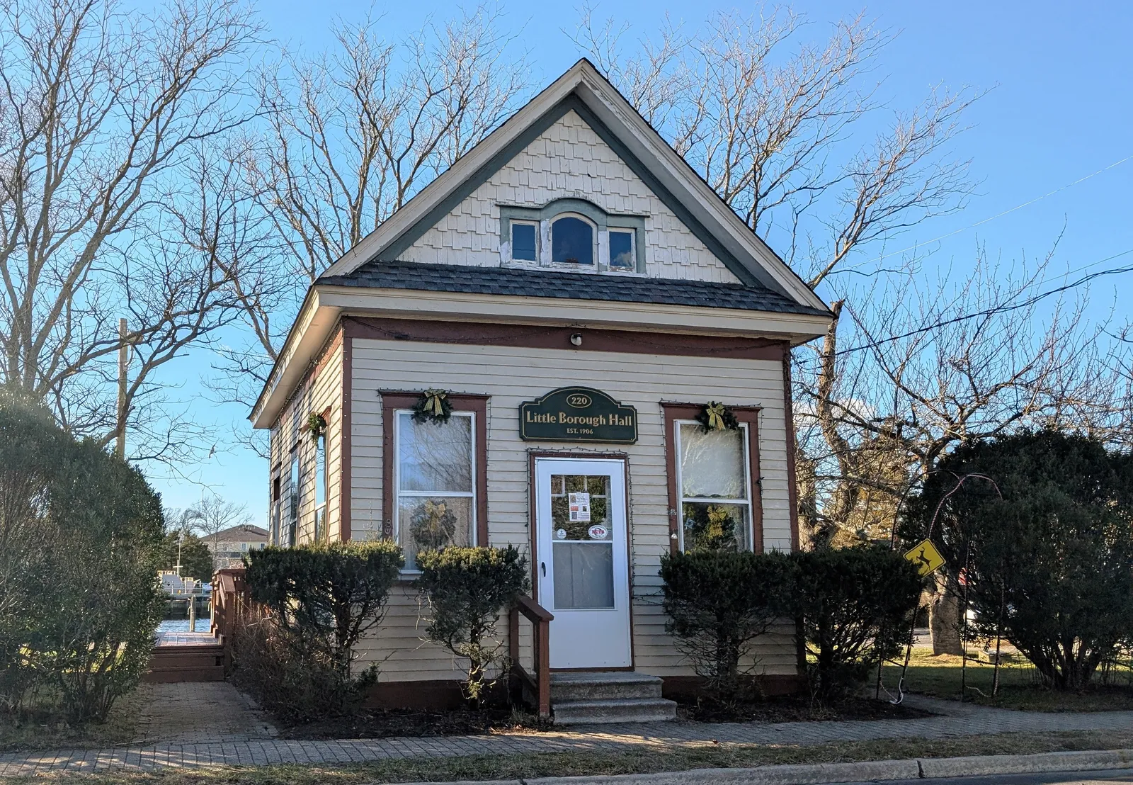 A small cream-colored historic building with a gabled roof, green sign and wreaths on the windows
