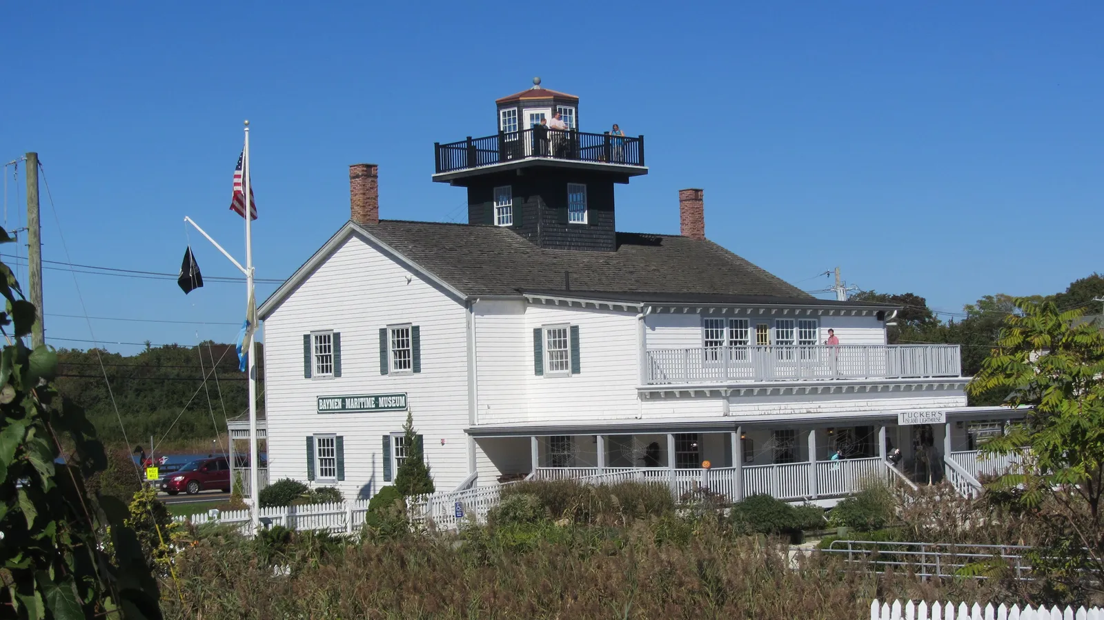 A white two-story lighthouse building with a black lantern room on the roof, flags flying beside it under a deep blue sky