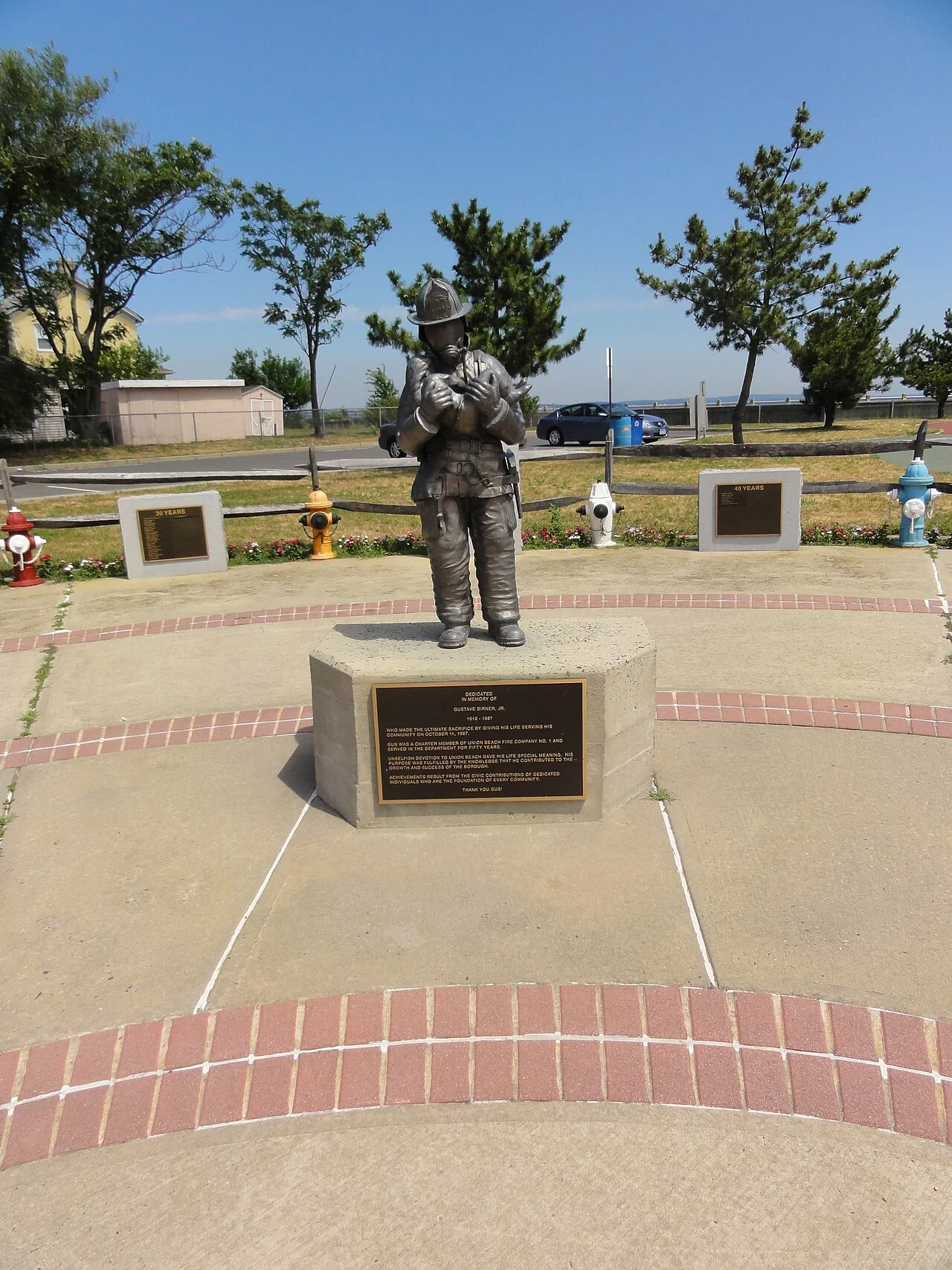 A bronze statue of a firefighter on a stone pedestal in a brick-edged plaza, with trees behind