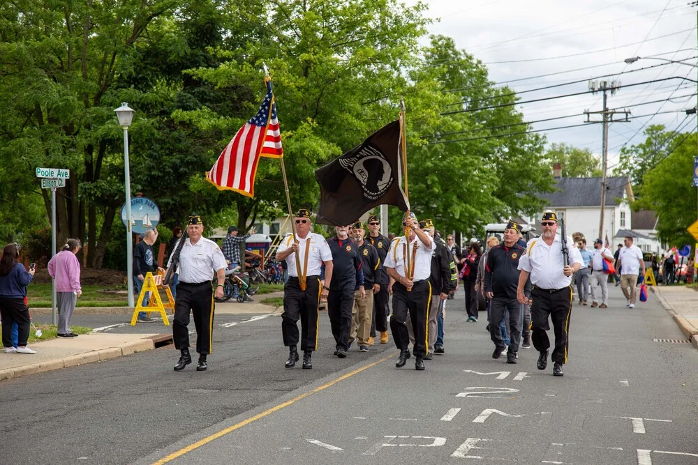 Veterans marching down a tree-lined street carrying the American flag and the POW-MIA flag, with spectators along the curb