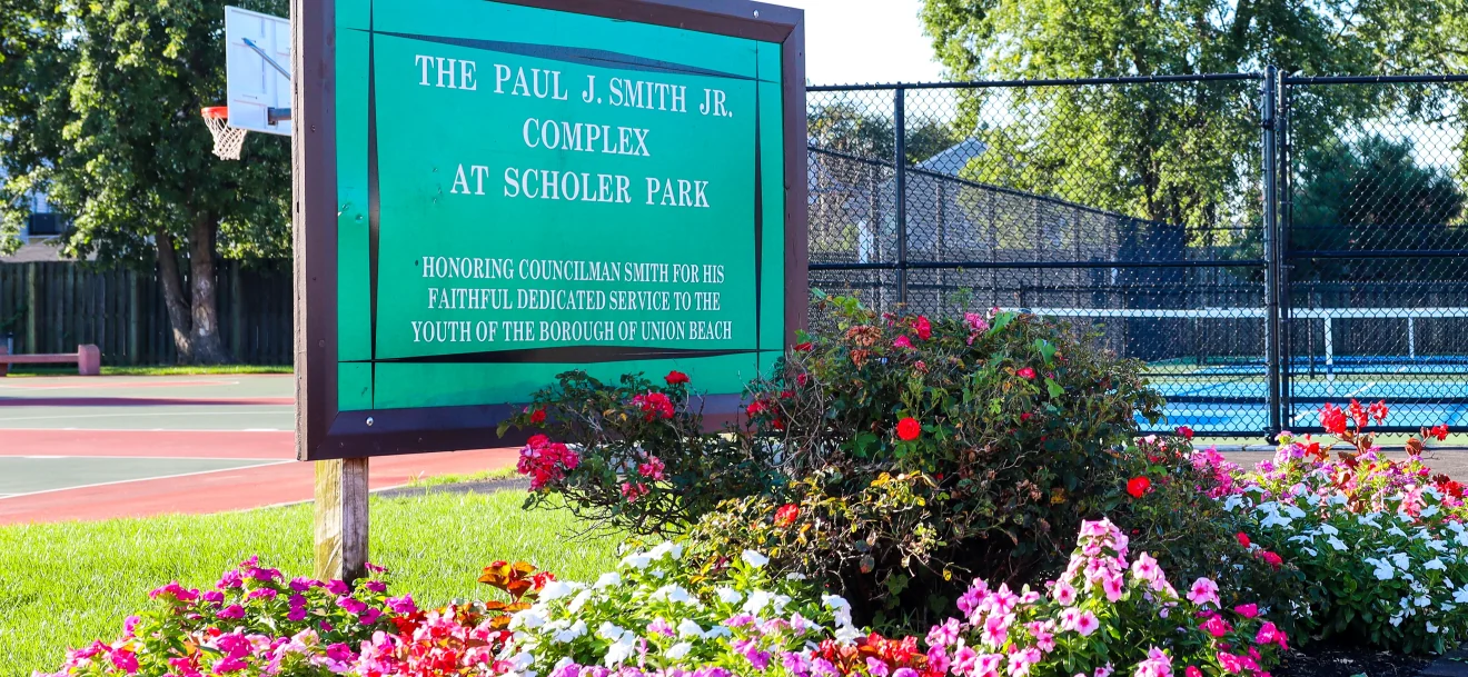 A green sign for the Paul J. Smith Jr. Complex at Scholer Park above a bed of pink and white flowers, with courts behind a fence