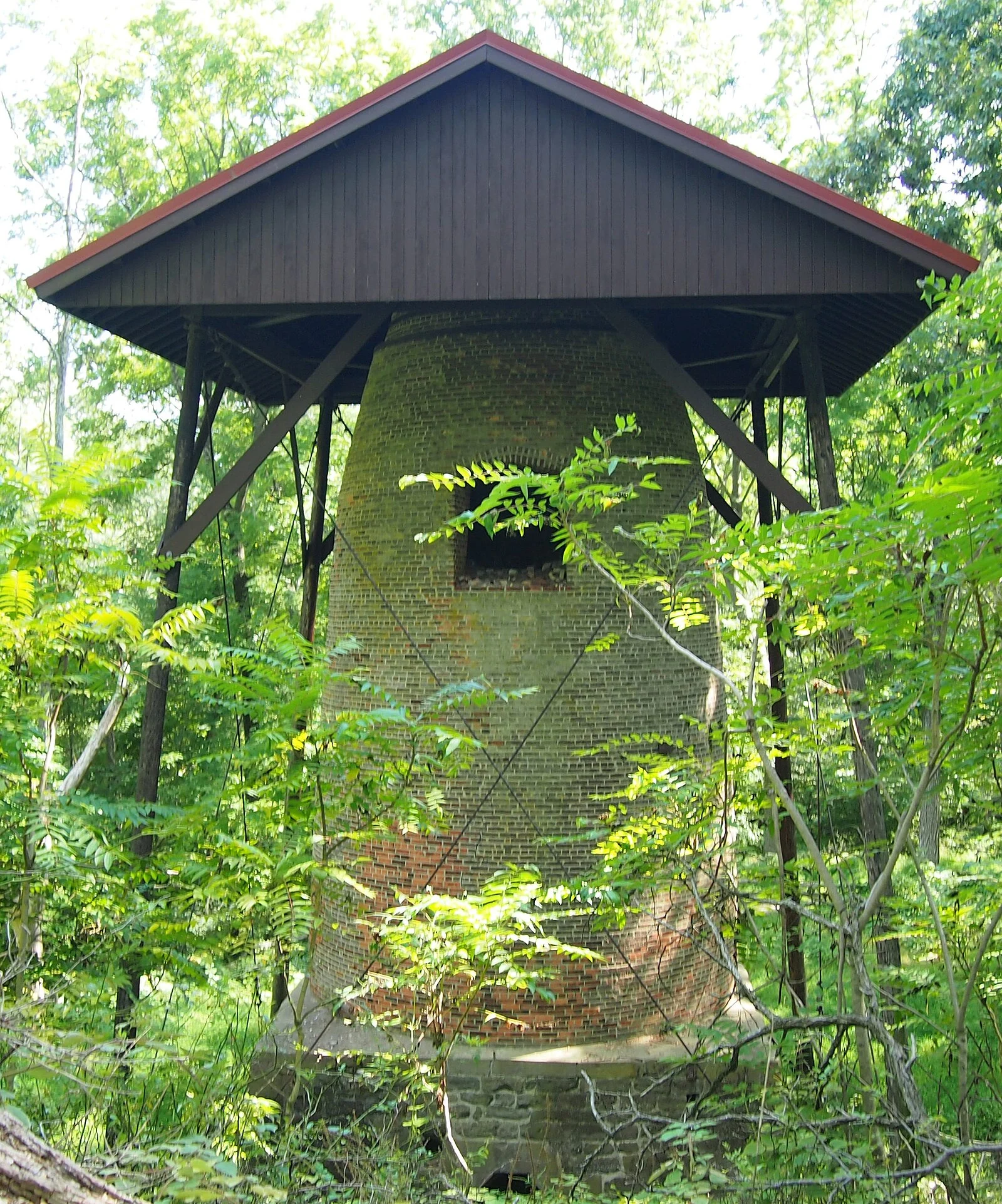 The tapered brick Howell Works blast furnace stack under a wooden shelter roof, surrounded by green woods
