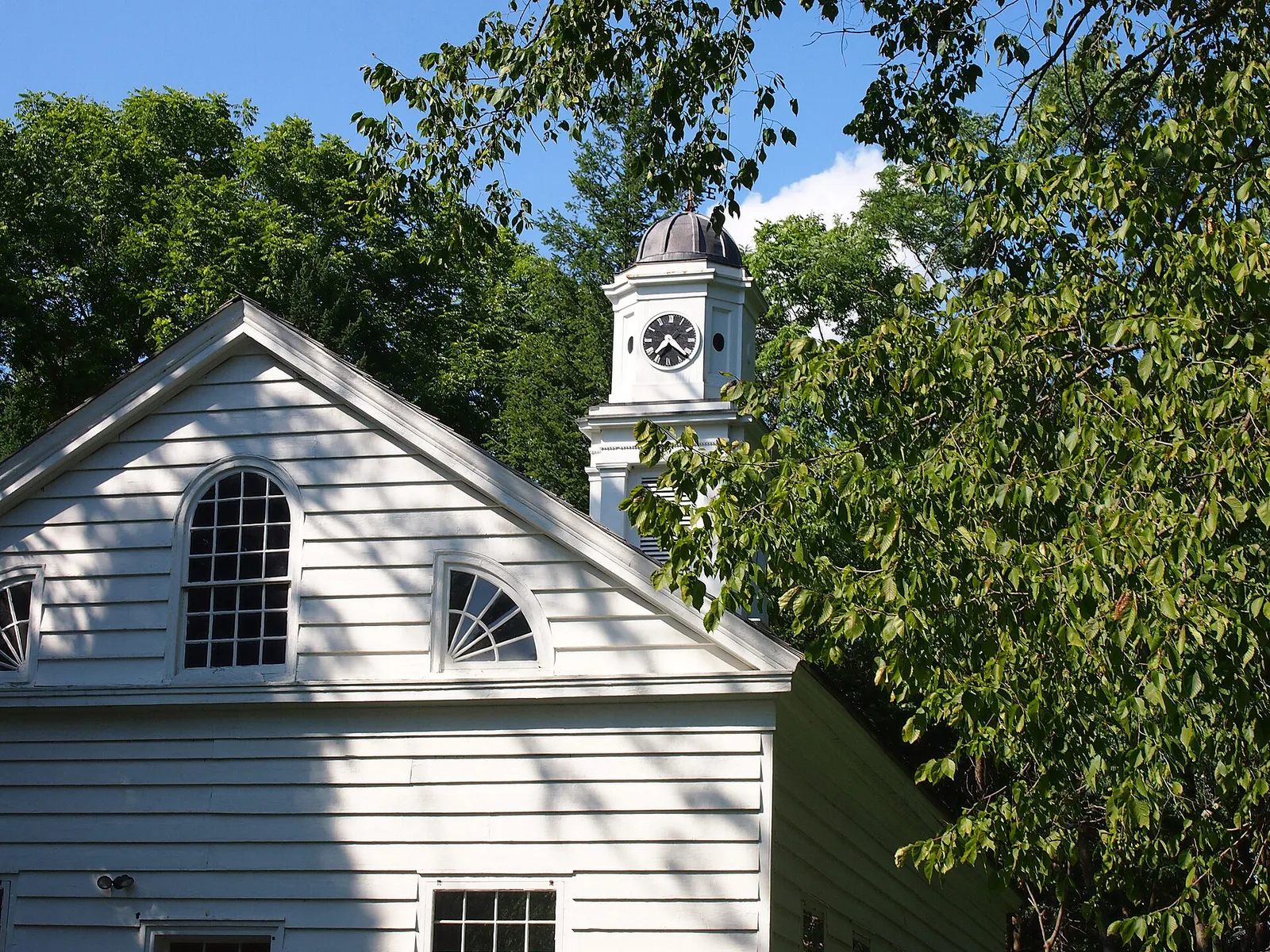 A white clapboard chapel gable with an arched window and a white clock tower against a blue sky and green trees