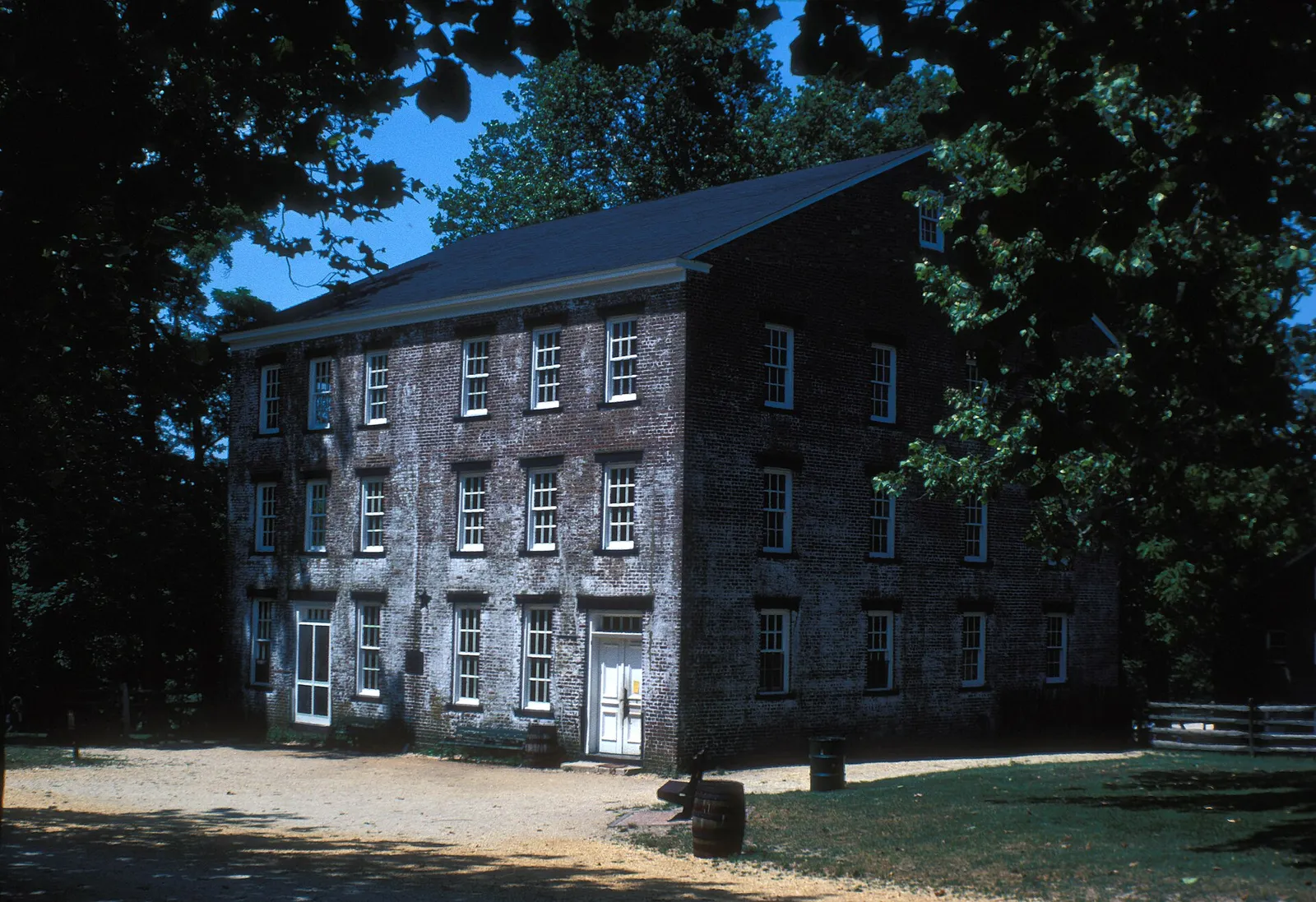 A four-story brick general store building with rows of white-trimmed windows in the shade of tall trees