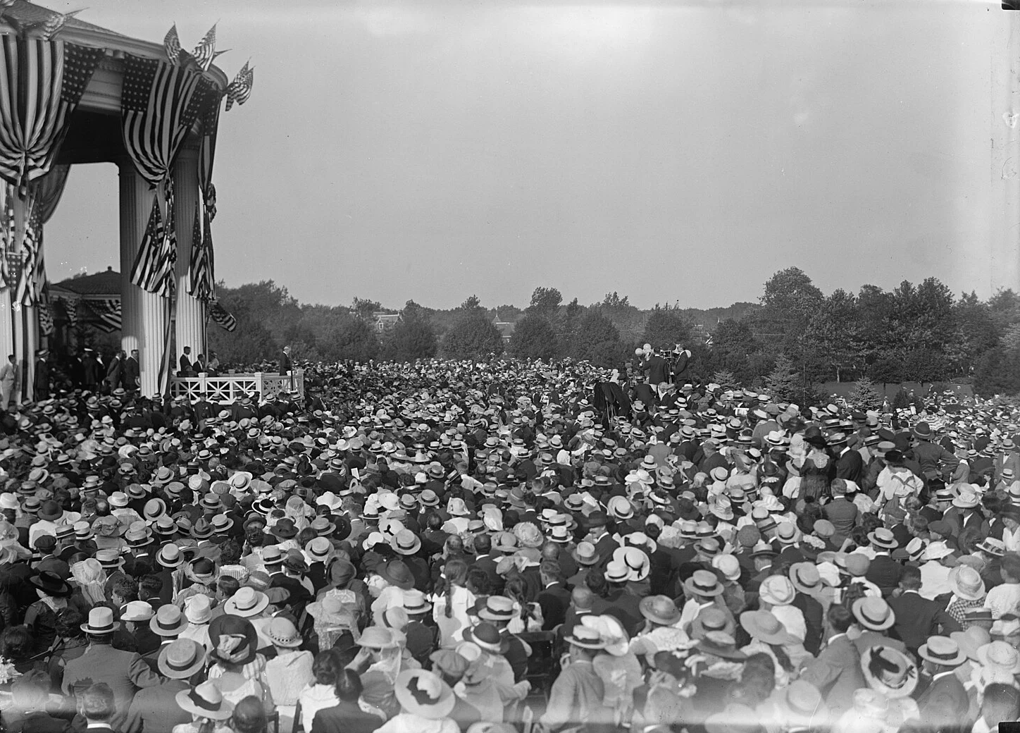A black-and-white photo of a vast crowd in straw hats on the lawn beside the flag-draped portico of the original Shadow Lawn