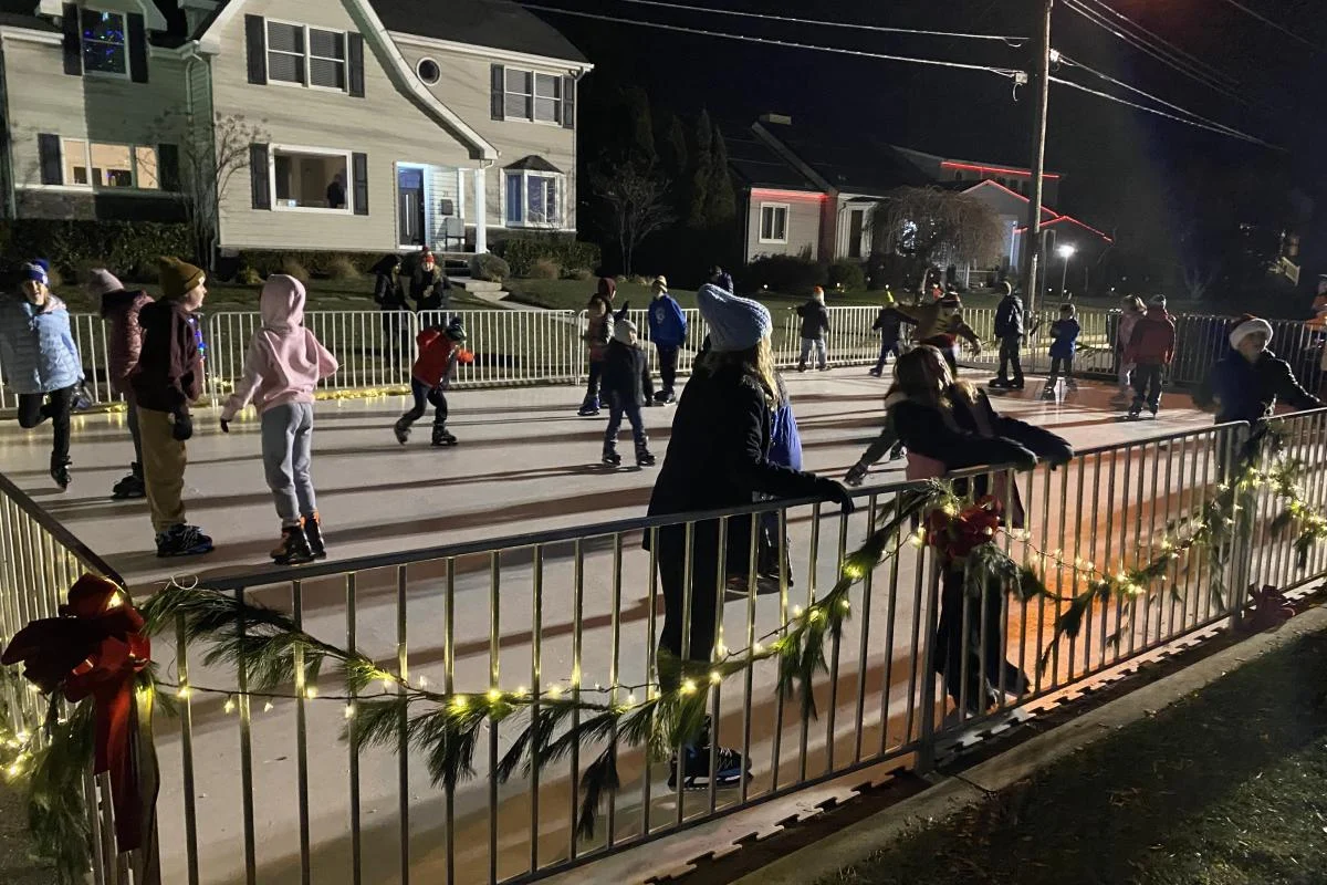 Skaters circling a small outdoor rink trimmed with garland and white lights on a December evening