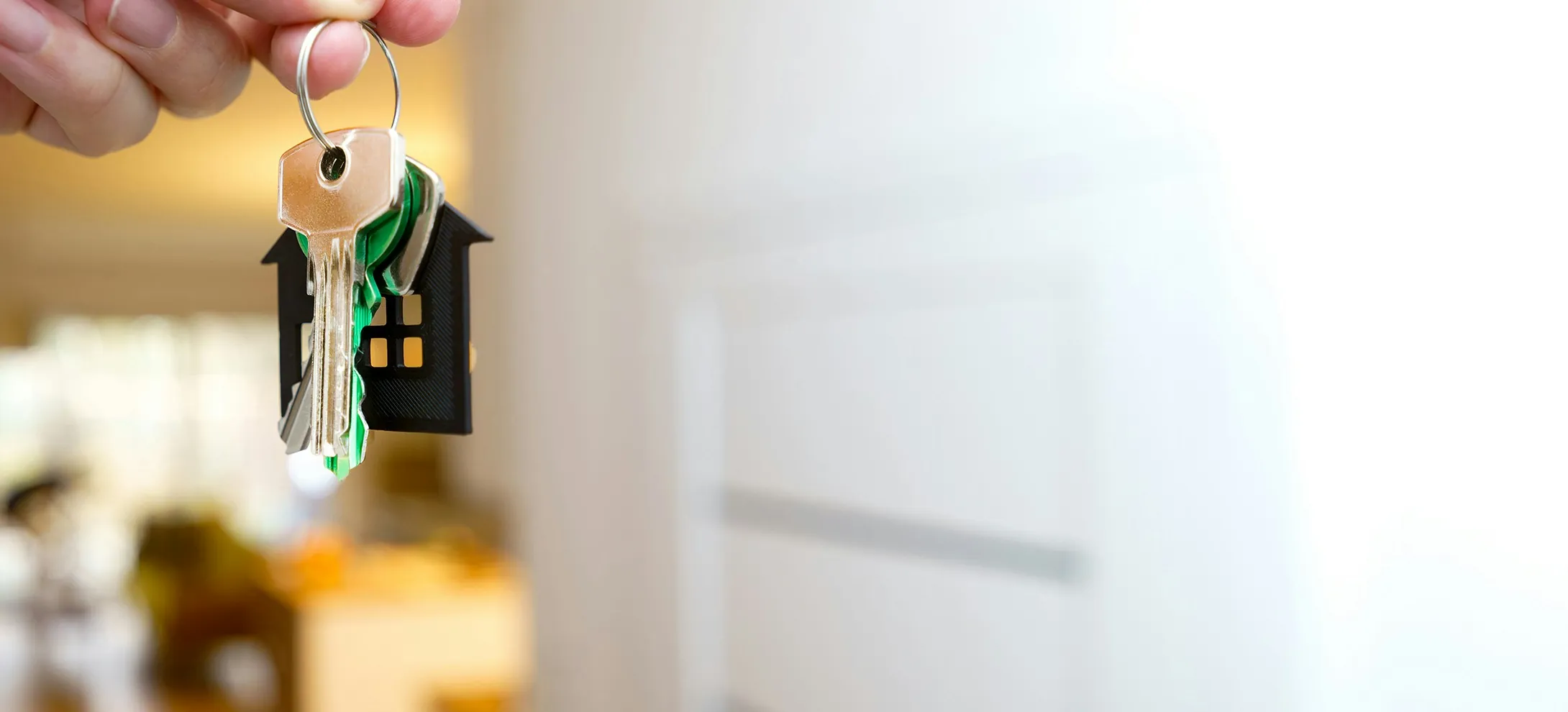 A hand holding a set of house keys on a small house-shaped key ring in a bright entryway
