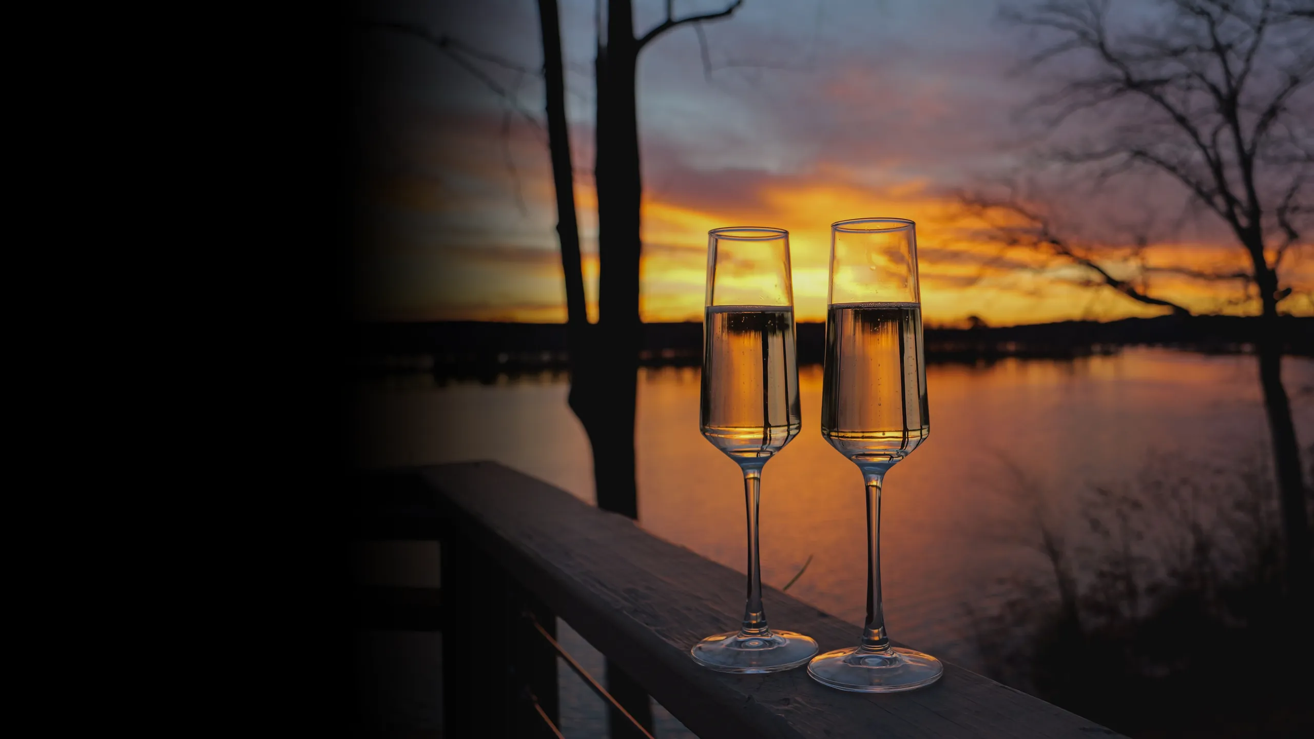 Two glasses of champagne on a deck railing above a lake at sunset