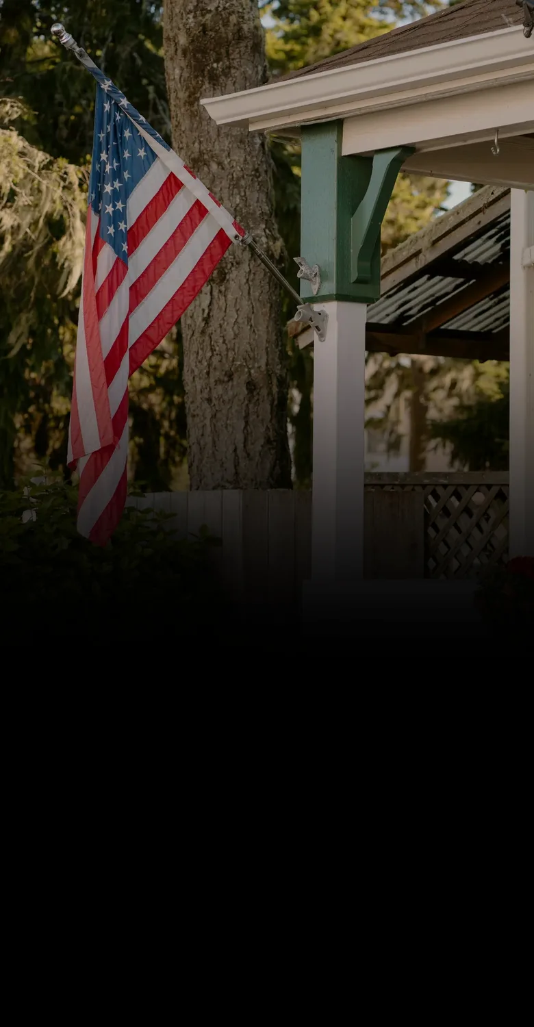 A white front porch with an American flag on its pole, fading into a fine-line drawing of a home beside a flag