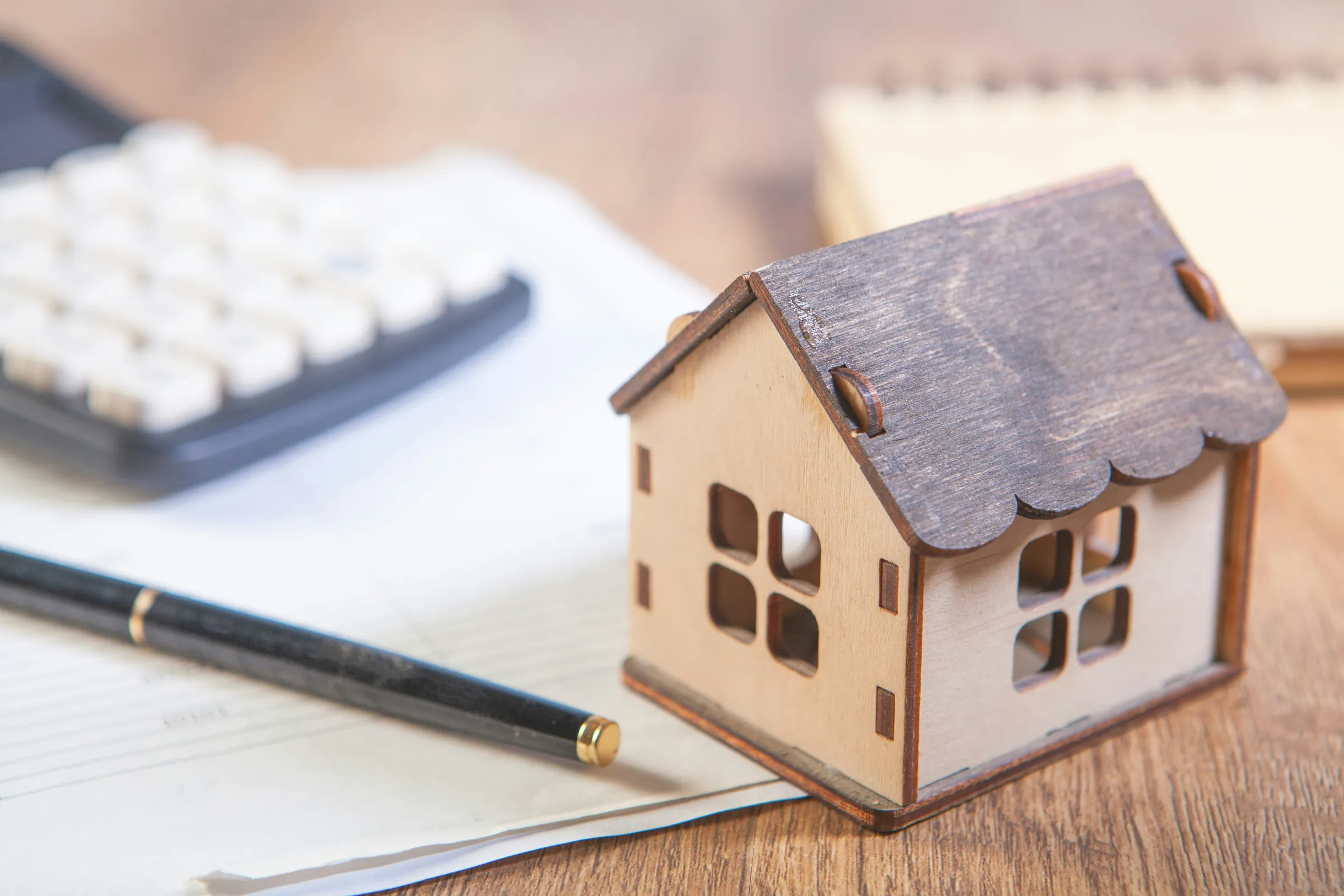 Small wooden model house beside a calculator, a pen and paperwork on a wooden desk