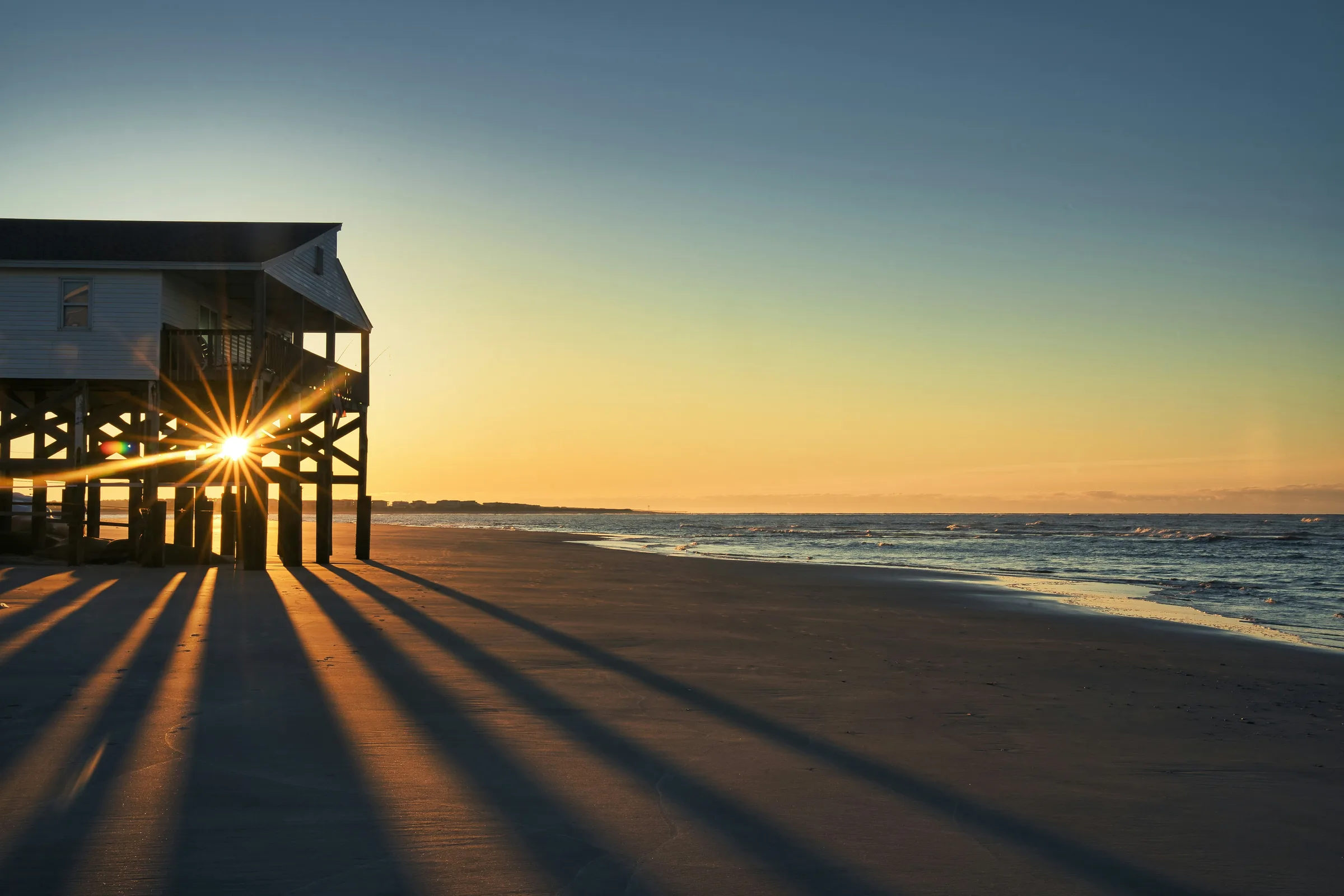 Beach house raised on tall wooden pilings, with the rising sun shining through the supports