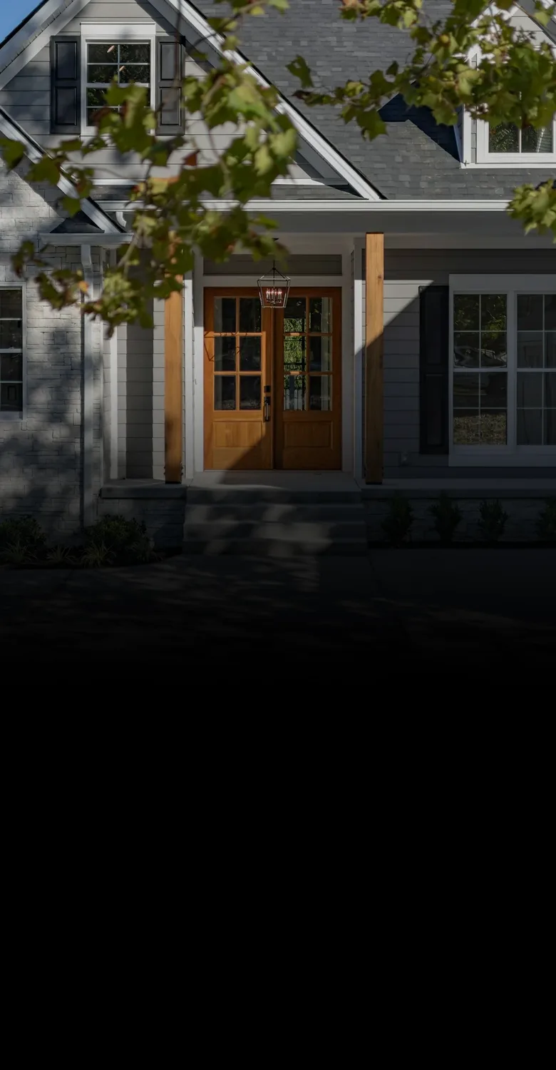 A suburban house with a stone front, gray siding and wooden front doors, fading into a fine-line drawing of a home with a red sign out front